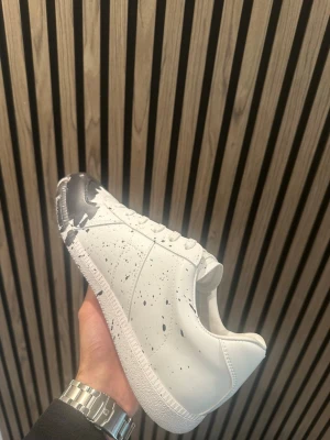 Vita sneakers maison  - Unika vita sneakers med svart färgstänk och svart tåparti. Skorna har lågt skaft, klassisk snörning och är tillverkade i slätt läder. Perfekt för dig som vill sticka ut med en kreativ och modern stil.