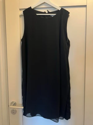 Svart ärmlös omlottklänning - Stilren svart ärmlös klänning med omlottdetalj framtill. Klänningen har en rak, enkel passform och är tillverkad i ett lätt, flytande material som ger ett elegant intryck. Perfekt för dig som gillar minimalistisk stil.
