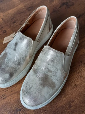 Metallic loafers från Ten Points storlek 37 - Snygga loafers i metallicfärgat läder från Ten Points. Skorna har en slip-on design med elastiska paneler på sidorna och vit sula. Insidan är fodrad i beige läder för extra komfort. Perfekta för dig som vill sticka ut med en unik look.