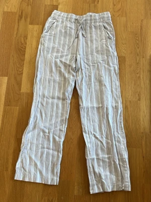 Beige randiga linnebyxor - Snygga beige linnebyxor med vita vertikala ränder och avslappnad passform. Byxorna har resår i midjan, fickor fram och bak samt raka ben. Perfekta för en chill och stilren look. Materialet känns lätt och luftigt, perfekt för varmare dagar.