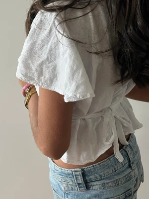 Vit omlottopp med knyt och kort ärm - Säljer en fräsch vit omlottopp med knyt i midjan och korta, vida ärmar. Toppen är croppad och har en lätt struktur i tyget, perfekt till jeans eller kjol. Enkel men ändå stylish design som passar till många looks.