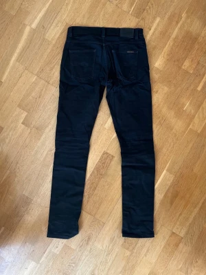 Svarta Dude Dan jeans från Nudie - Snygga svarta slim fit jeans från Nudie Jeans med klassisk femficksdesign och normal midja. Jeansen har raka ben och diskreta detaljer, perfekta för en stilren look. Materialet är stretchigt för extra komfort. Modell är Dude Dan och storleken är 32/36. Tyvärr hål i skrevet, går att laga enkelt eller vid extra begär kan jag lösa det.