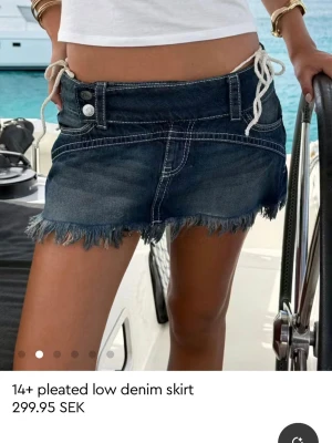 Mörkblå denim minikjol med snörning - Trendig mörkblå jeanskjol i kort modell med rå fransad kant och vita snörningsdetaljer i midjan. Kjolen har dubbla knappar och markerade sömmar för en cool look. Perfekt för dig som gillar streetstyle och denim.