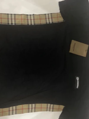 Svart t-shirt med rutiga detaljer - Snygg svart t-shirt från Burberry med klassiska beige-rutiga detaljer vid ärmslut och halsringning. T-shirten har en enkel design med korta ärmar och diskret Burberry-logga på bröstet. Perfekt för en stilren och trendig look.