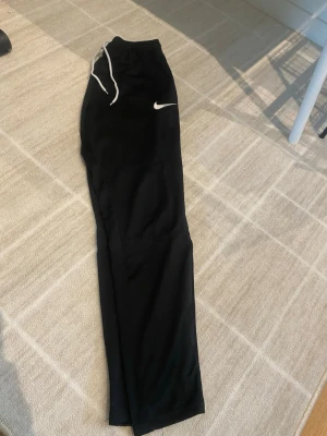 Svarta Nike träningsbyxor - Säljer ett par svarta träningsbyxor från Nike med vit logga på höften och vita dragsnören i midjan. Byxorna har en rak passform och är tillverkade i ett lätt och bekvämt material som passar perfekt till träning. Ej tights utan mer mjukis 