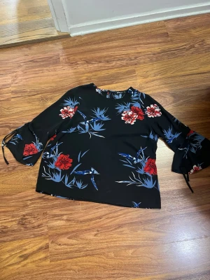 Svart blommig blus med vida ärmar - Svart blus med röda och blå blommor, vida trekvartsärmar med snörning vid ärmslut. Blusen har en rak passform och rund halsringning. Perfekt för dig som vill ha en färgstark och trendig look.