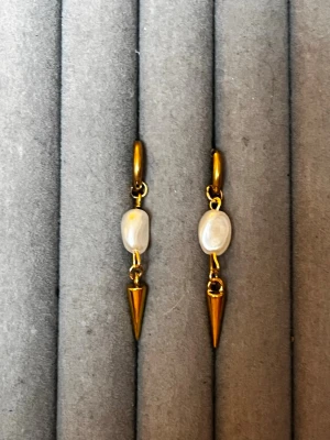 Guldiga örhängen med pärlor och spikes - Snygga örhängen i guldton med oregelbundna vita pärlor och spetsiga hängen. Perfekt accessoar för dig som gillar en mix av klassiskt och edgy stil. Örhängena har små ringar upptill och är tillverkade i metall med pärldetaljer.
