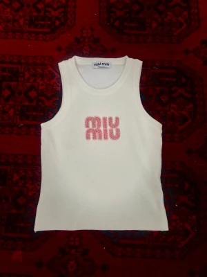 Vit tanktop från Miu Miu - Säljer en vit ärmlös topp från Miu Miu med rosa logga broderad framtill. Perfekt att styla med jeans eller kjol för en clean och trendig look. Materialet känns mjukt och följsamt.