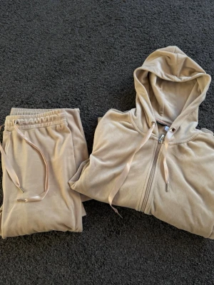 Beige velour loungewear-set med huva - Mysigt loungewear-set i beige velour med matchande byxor och hoodie. Hoodien har dragkedja och snörning i huvan, medan byxorna har resårmidja och snören. Perfekt för chill dagar hemma. Olika storlekar!! Tröjan M och byxorna xs 