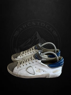 Philippe Models - Snygga vita sneakers från Philippe Model med blå hälkappa och grå detaljer vid tån. Klassisk låg modell med perforerade sidor och ikonisk sköld på sidan. Skorna har vita snören och en robust gummisula. Perfekta för en stilren streetlook.