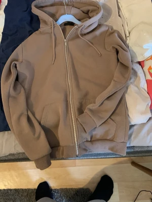 Beige ziphoodie med huva - En stilren beige hoodie med dragkedja och huva. Modellen har ribbade muddar vid ärmslut och nederkant samt två fickor framtill. Perfekt för en avslappnad look. Materialet känns mjukt och bekvämt, troligen bomullsblandning.