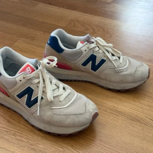 New Balance sneakers beige/blå/röd - Snygga sneakers från New Balance i beige textil och mocka med blå detaljer och röd häl. Klassisk N-logga på sidan och vit sula. Sköna och stilrena. Storlek 42,5 (EU). Använda 1 säsong, men har problem med fötterna så kan inte ha snygg-skor. Inköpta för 1.000 kr förra sommaren.