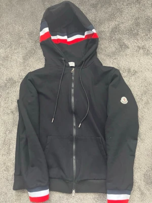 Svart Moncler hoodie med dragkedja - Svart hoodie från Moncler med hel dragkedja, huva med snören och röd, vit och blå rand på huvan. Ärmsluten har matchande ränder och vänster ärm har Moncler-logga. Tillverkad i mjukt bomullsmaterial och har två fickor framtill.