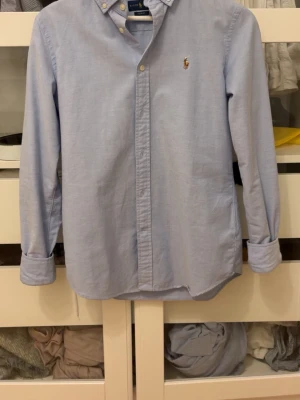 Ljusblå skjorta från Ralph Lauren - Klassisk ljusblå skjorta från Ralph Lauren med broderad logga på bröstet. Skjortan har knappar framtill, button-down krage och långa ärmar. Perfekt för en stilren och tidlös look. Bara att skriva vid vidare frågor.