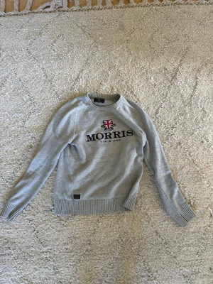 Grå tröja från Morris med logga - Snygg grå tröja från Morris med broderad logga och brittisk flagga på bröstet. Tröjan har rund halsringning, ribbade muddar och är tillverkad i mjukt stickat material. Perfekt för en stilren och avslappnad look. Storlek S inga defekter eller skador.