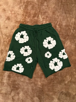 Gröna shorts från Denim Tears M - Mörkgröna shorts från Denim Tears med vita blommönster över hela plagget. De har resår i midjan och dragsko för justerbar passform. Perfekta för en avslappnad och trendig stil.