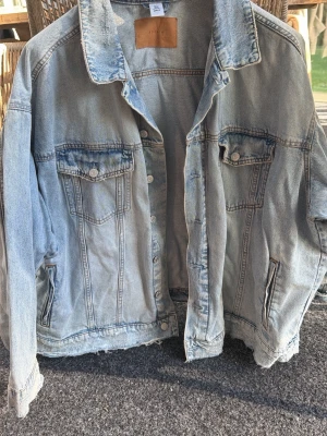 Ljusblå jeansjacka från &Denim XXL - Snygg ljusblå jeansjacka från &Denim i storlek XXL. Klassisk modell med två bröstfickor med lock och knappar samt sidofickor. Jackan har en avslappnad passform och slitna detaljer vid fållen. Sparsamt använd. 