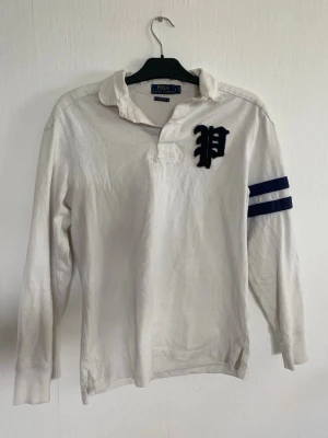 Polo Ralph Lauren rugbytröja  - Vit långärmad henley-skjorta från Polo Ralph Lauren med blått P-tryck på bröstet och två blå ränder på ena ärmen. Tröjan har krage, knappar upptill och är gjord i bomull för en bekväm känsla. Perfekt för en avslappnad men stilig outfit 🌞