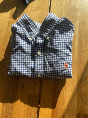 Blårutig skjorta från Ralph Lauren - Klassisk blårutig skjorta från Ralph Lauren med vit knappstängning och broderad logga i orange på bröstet. Skjortan har en tidlös krage och är tillverkad i mjuk bomull. Perfekt för en stilren look.