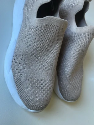 Beige slip-on sneakers med stickad ovandel stl 34 - Snygga beige slip-on sneakers stl 34 med stickad ovandel och vit chunky sula. Skorna har en modern, minimalistisk design utan snörning och är tillverkade i ett mjukt, ventilerande textilmaterial som ger en skön passform. Perfekta för en avslappnad och trendig stil.