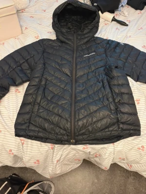 Svart pufferjacka med huva - Svart pufferjacka från Peak Performance med huva och dragkedja framtill. Jackan har ett quiltat mönster i V-form och är vadderad för extra värme. Perfekt för kalla dagar och har en stilren design med diskret logga på bröstet. Litet hål i armen 