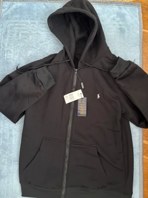 Svart hoodie från Polo Ralph Lauren - Svart hoodie med dragkedja från Polo Ralph Lauren. Klassisk design med huva, dragsko och två stora fickor framtill. Liten vit logga broderad på bröstet. Tillverkad i mjukt bomullsmaterial för en skön och avslappnad stil.
