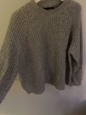 Grå stickad tröja från 157 - Mysig grå stickad tröja från 157 med grovstickat mönster och rund halsringning. Tröjan har långa ärmar och ribbade muddar vid hals, ärmslut och nederkant. Perfekt för kyliga dagar och enkel att matcha med olika outfits.