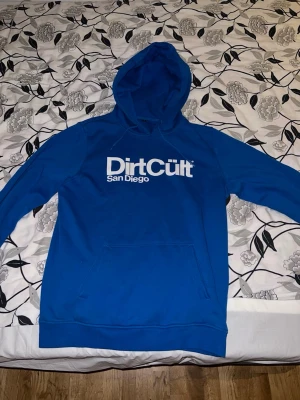 Blå DirtCült hoodie med tryck - Snygg blå hoodie från DirtCült med stort vitt tryck 'DirtCült San Diego' på bröstet. Klassisk modell med huva och känguruficka framtill. Perfekt för dig som gillar streetwear och vill sticka ut med en färgstark look. Använd 2-3 gånger. Storlek L men skulle passa M också då jag har M storlek och den passar mig