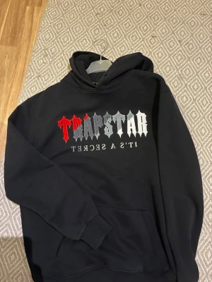 Svart Trapstar hoodie med tryck - Säljer en svart hoodie från Trapstar med stor logga i rött och vitt på bröstet och texten 'IT'S A SECRET' under. Klassisk passform med huva och känguruficka framtill. Perfekt för dig som gillar streetwear och vill sticka ut med en fet design.