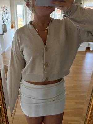 Beige cropped kofta med knappar - Trendig beige cropped kofta med stora knappar framtill och v-ringad hals. Koftan har långa ärmar och är tillverkad i ett tunt, stickat material som känns mjukt mot huden. Perfekt att styla med kjol eller jeans för en snygg look.