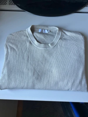 Beige stickad tröja från Ljung - Snygg beige ribbstickad tröja från Ljung by Marcus Larsson. Tröjan har rund halsringning och är tillverkad i ett mjukt, stickat material som känns skönt mot huden. Perfekt för dig som gillar stilrena och enkla plagg med modern känsla.