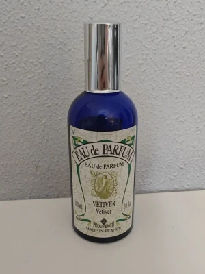 Eau de Parfum Vetiver från Provenco - Eau de Parfum Vetiver från Provenco i en elegant blå glasflaska med silverfärgad spraykork. Flaskan rymmer 100 ml och har en klassisk etikett med gröna detaljer. Parfymen är tillverkad i Frankrike. Uppskattad mängd kvar 60%, se bild för markering. 