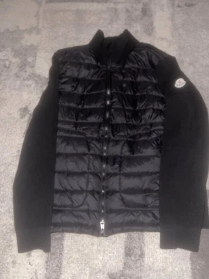 Svart cardiganjacka från Moncler - Snygg svart cardiganjacka från Moncler med quiltad front och stickade ärmar. Jackan har dragkedja och Moncler-logga på ena ärmen. Perfekt för lager-på-lager och ger en cool look. Materialet är en mix av nylon och stickad ull. Pris kan diskuteras vid snabb affär!
