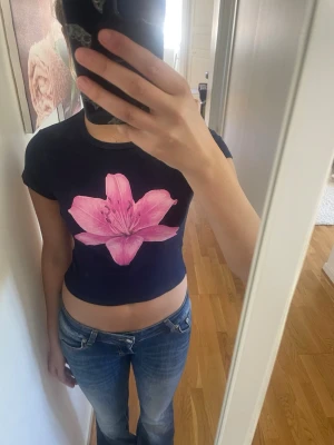 Mörkblå croppad t-shirt med blomma newyorker - Säljer en mörkblå croppad t-shirt med en stor rosa blomma på bröstet. T-shirten har korta ärmar och är gjord i mjuk bomull. Perfekt för dig som vill ha en cool och färgstark look.