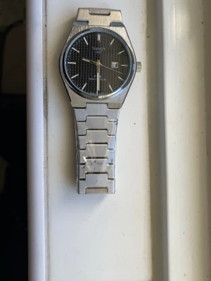 Tissot PRX Powermatic 80 klocka - Snygg Tissot PRX Powermatic 80 klocka med svart urtavla och datumfönster. Klockan har ett stilrent rutmönster på tavlan och ett armband i silverfärgad metall. Perfekt accessoar för dig som gillar klassisk och modern design.