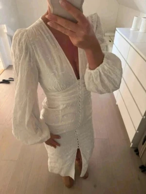 Vit långklänning från Gina Tricot - Superfin vit långklänning från Gina Tricot med v-ringning, puffiga långärmar och knappar framtill. Klänningen har broderade blommor i tyget och en slits framtill. Perfekt för sommaren! Endast använd 1 gång!