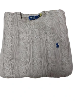 Beige kabelstickad tröja Polo Ralph Lauren - Kabelstickad beige tröja från Polo Ralph Lauren med rund halsringning och klassisk blå broderad logga på bröstet. Tröjan är i mjukt material och har ribbade muddar vid hals, ärmslut och nederkant. Perfekt för en stilren och avslappnad look.