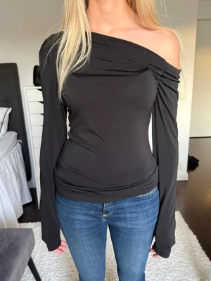 Svart offshoulder topp - Trendig svart offshoulder topp med långa ärmar och asymmetrisk halsringning. Sitter tight och har en snygg drapering över axeln. 