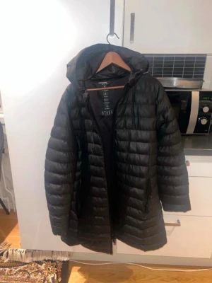 Svart lång dunjacka från Etage - Säljer en svart lång dunjacka från Etage med huva och dragkedja framtill. Jackan har två sidofickor med dragkedja och quiltad design som ger en snygg look. Perfekt för kalla vinterdagar och riktigt varm tack vare dunfyllningen.