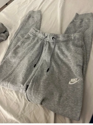 Grått Nike mjukisset med hoodie - Säljer ett grått mjukisset från Nike med matchande hoodie och byxor. Hoodien har dragsko och magficka, byxorna har resår och dragsko i midjan samt sidofickor. Båda plaggen har Nike-logga framtill. Perfekt för chill dagar.
