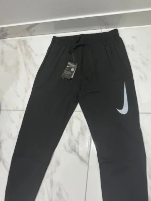 Svarta Nike mjukisbyxor med logga - Svarta mjukisbyxor från Nike med stor vit Swoosh-logga på benet. Byxorna har resår i midjan med dragsko och sidofickor. Perfekta för chill eller träning, tillverkade i mjukt och bekvämt material.