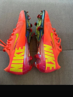 Nike Mercurial fotbollsskor neonröda - Säljer ett par Nike Mercurial fotbollsskor i en riktigt snygg neonröd färg med stora gula NIKE-loggor på sidorna. Skorna har snörning, låg profil och en cool, skimrande yttersula med dobbar för bästa grepp på planen. Perfekta för dig som vill sticka ut på fotbollsplanen.