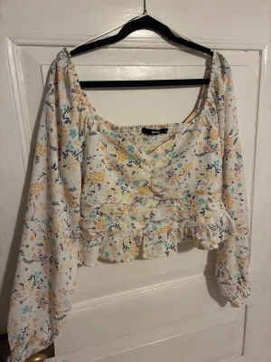 Blommig blus från Bik Bok med volang - Supersöt blommig blus från Bik Bok i vitt med gula, blå och lila blommor. Blusen har långärmade puffärmar, v-ringning och volangkant nedtill. Perfekt för dig som gillar romantisk och trendig stil. Tunn och luftig i materialet, troligtvis polyester.