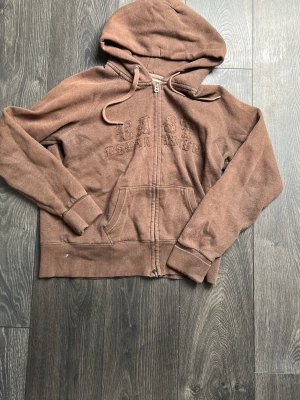 Brun zip-hoodie med tryck - Säljer en brun hoodie med dragkedja från East Department. Hoodien har huva med dragsko, känguruficka och broderat tryck på bröstet. Perfekt för en chill och avslappnad stil. Materialet är mjukt och skönt, troligen bomullsblandning. Kan gå ner u pris vid snabb affär.