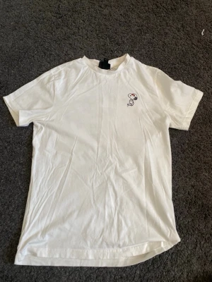 Vit Snoopy t-shirt med graffiti-tryck - Vit t-shirt med coolt graffiti-inspirerat tryck av Snoopy och texten 'Big Deal' på ryggen. Liten Snoopy-detalj framtill. Skön passform och klassisk rund hals. Perfekt för dig som gillar streetstyle och vill sticka ut lite extra. Från h&m o storlek small