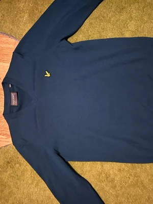 Mörkblå tröja från Lyle & Scott - Mörkblå långärmad tröja från Lyle & Scott med V-ringning och klassisk gul logga broderad på bröstet. Enkel och stilren design som passar perfekt till jeans eller chinos.