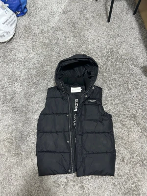 Svart dunväst från Calvin Klein Jeans - Snygg svart dunväst från Calvin Klein Jeans med huva och diskret logga på bröstet. Västen har quiltad design, dragkedja och tryckknappar framtill samt en praktisk bröstficka. Perfekt för lager-på-lager och streetstyle.