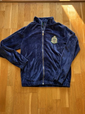Blå ziptröja i fleece från New Black - Mjuk och skön blå ziptröja i fleece från New Black med matchande byxor till. Tröjan har hel dragkedja, ståkrage och broderad patch med glob och krona på bröstet. Perfekt för lager-på-lager och chill dagar.