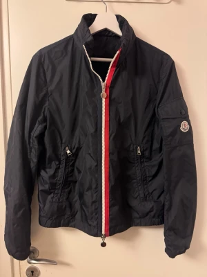 Svart vindjacka från Moncler - Snygg svart vindjacka från Moncler med vit och röd dragkedja framtill. Jackan har ståkrage, två fickor med dragkedja och Moncler-logga på ärmen. Tillverkad i lätt och glansig nylon, perfekt för blåsiga dagar.Moncler Keralle Windbreaker. Colour: Navy. Size: 2(S/M). Very good condition.