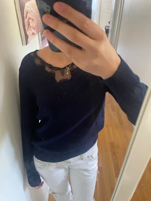 Mörkblå tröja med spetsdetalj - Snygg mörkblå tröja med lång ärm och unik spetsdetalj vid halsringningen. Tröjan har en rundad hals och ribbade muddar vid ärmslut och nederkant. Perfekt för dig som gillar stilrena plagg med en twist.